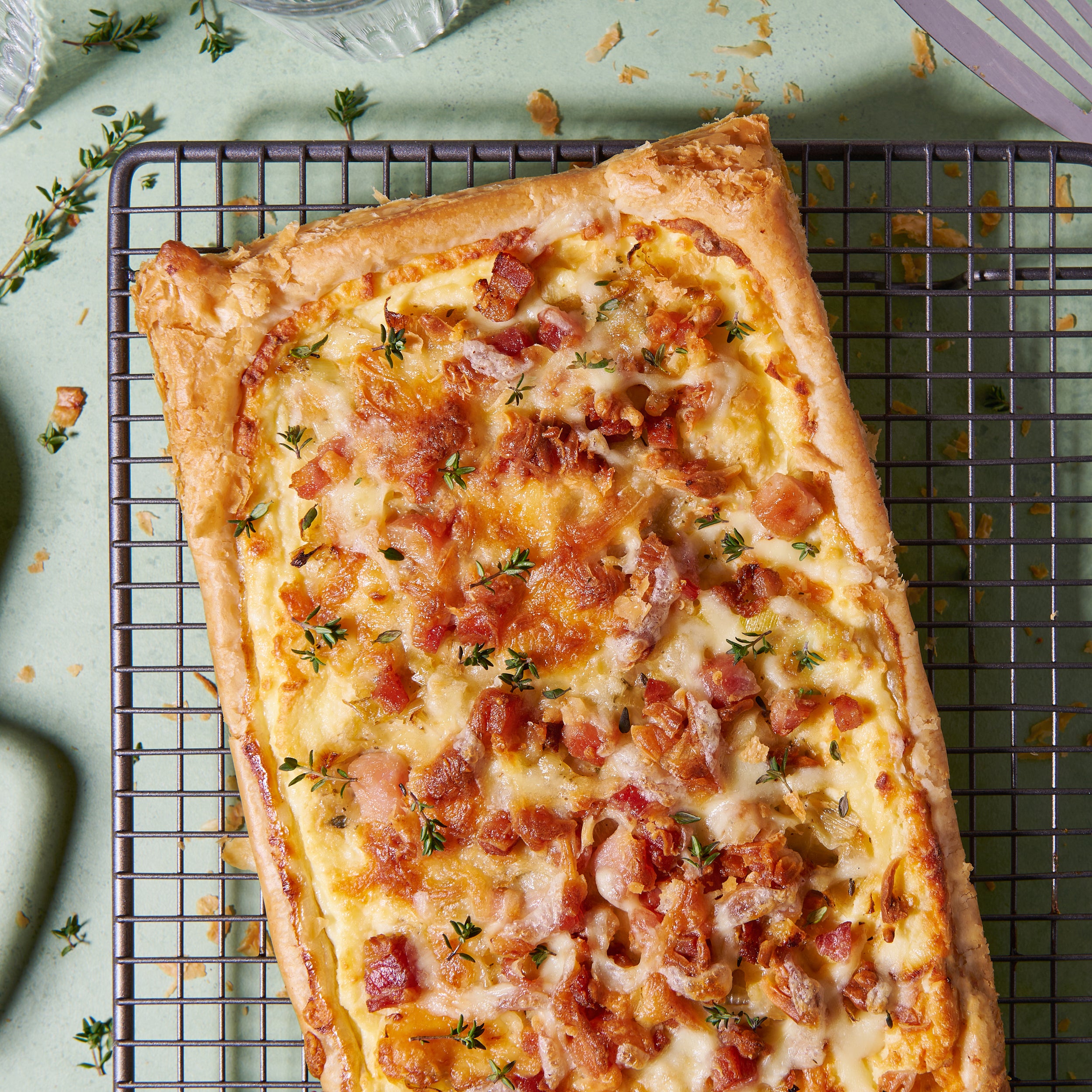 Bacon, leek and ricotta tart with a golden puff pastry crust, topped with melted cheese and fresh thyme, on a cooling rack