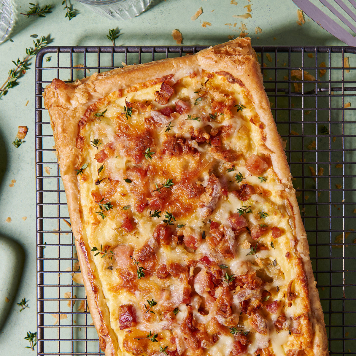 Bacon, leek and ricotta tart with a golden puff pastry crust, topped with melted cheese and fresh thyme, on a cooling rack