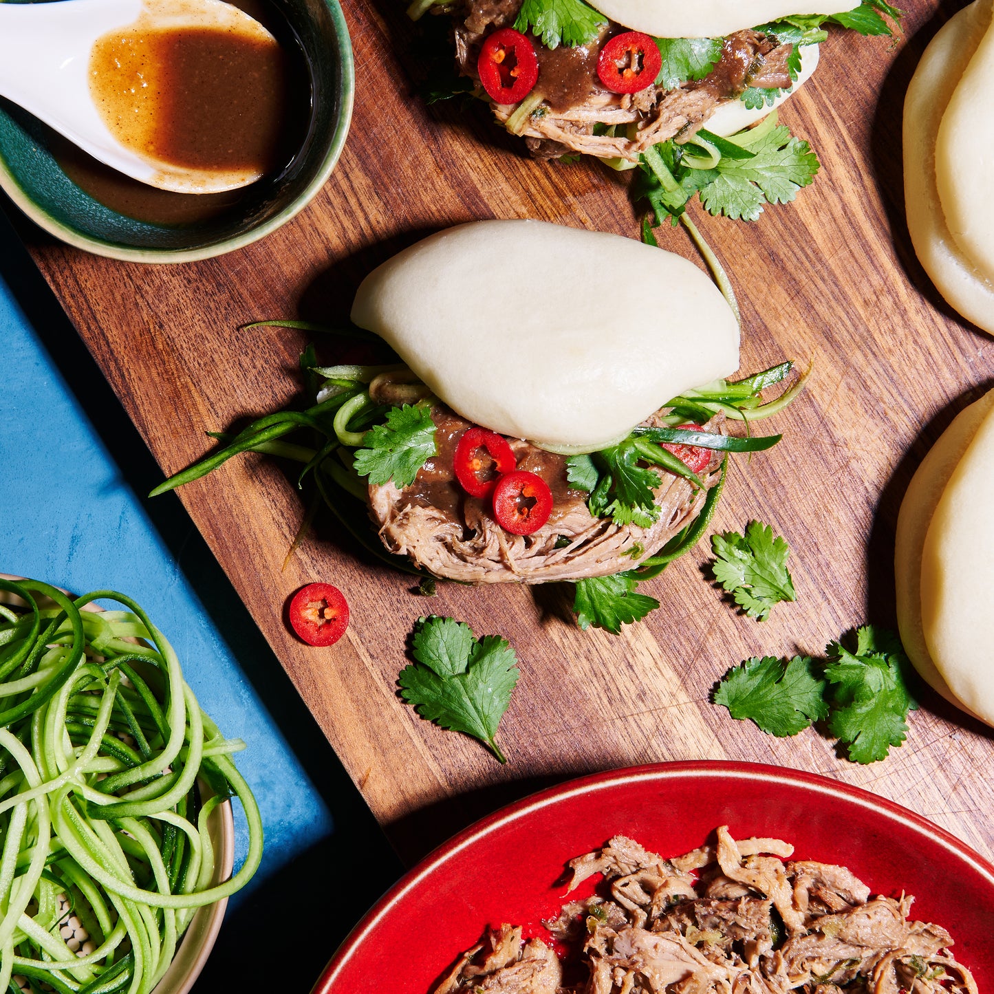 Overhead view of bao buns filled with shredded hoisin pork, coriander, red chilli, and cucumber on a wooden board with dipping sauce.
