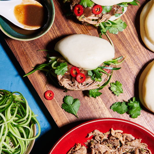 Overhead view of bao buns filled with shredded hoisin pork, coriander, red chilli, and cucumber on a wooden board with dipping sauce.
