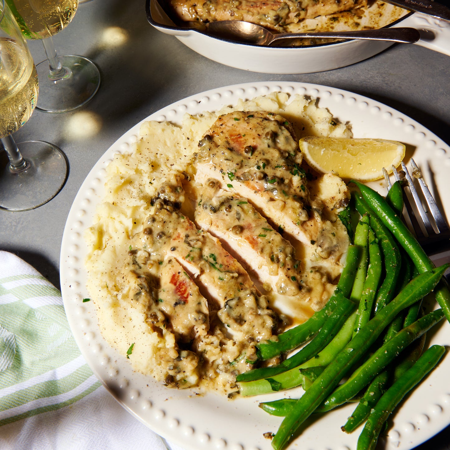 Sliced chicken scallopine with green peppercorn sauce plated over mashed potato, served with green beans and a lemon wedge