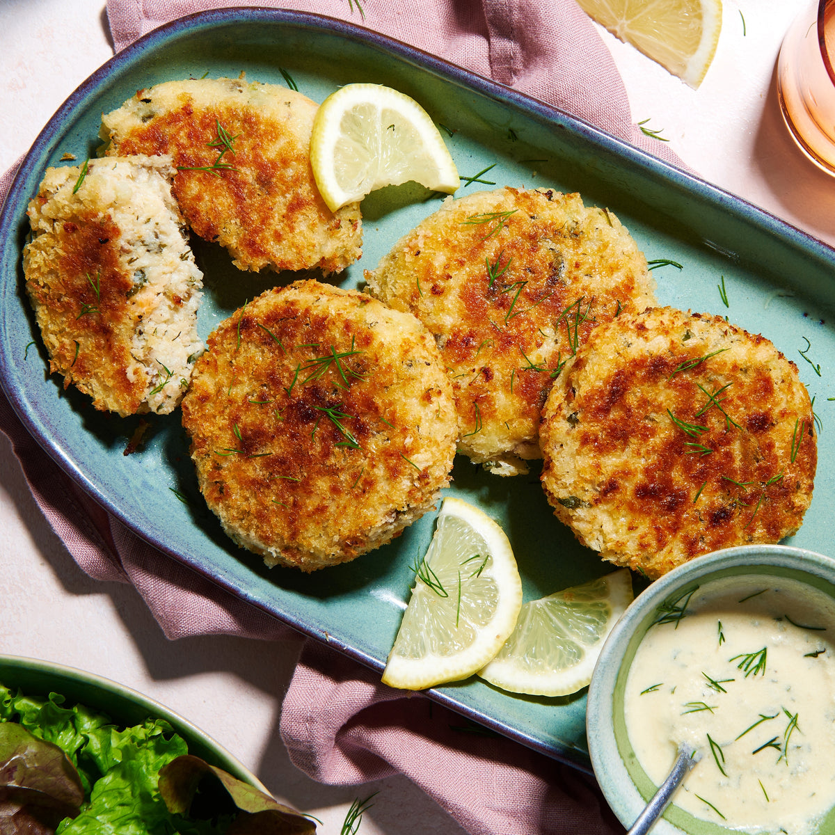 Plate of golden Salmon & Barramundi Fish Cakes with lemon wedges, fresh dill, and a creamy dipping sauce.
