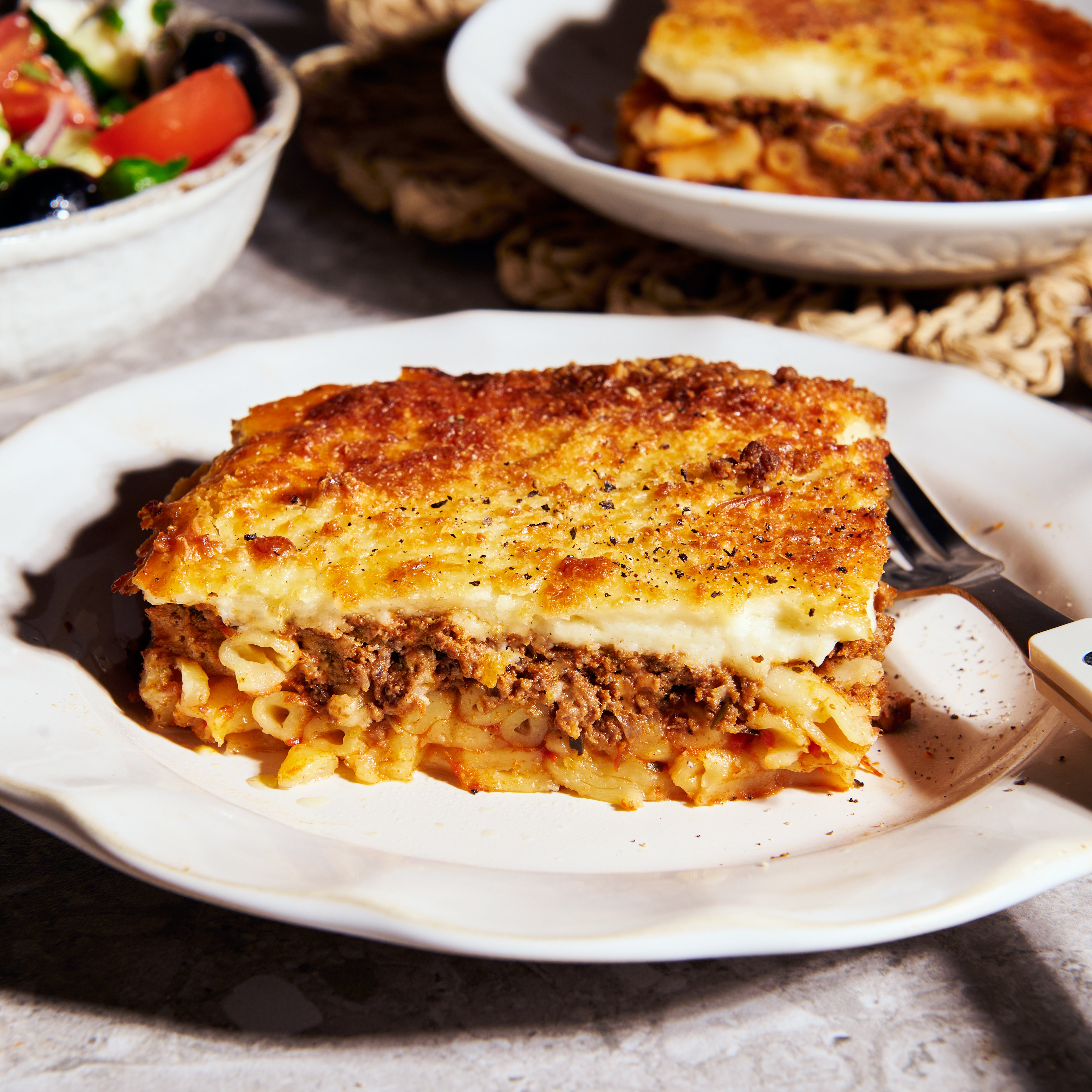 Slice of lamb pastitsio plated, layered pasta and lamb topped with a golden baked bechamel crust, served with a Greek salad