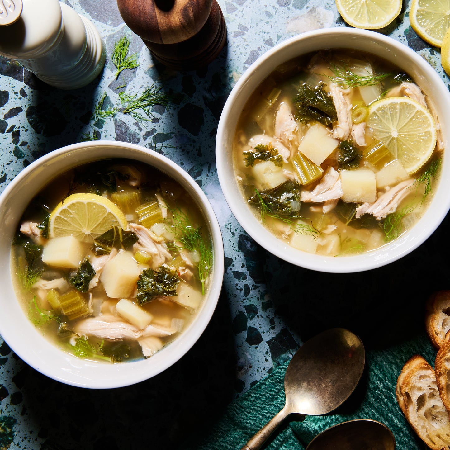 Overhead view of two bowls of Lemony Chicken Soup with fennel, potatoes, kale, and lemon slices on a blue speckled surface.
