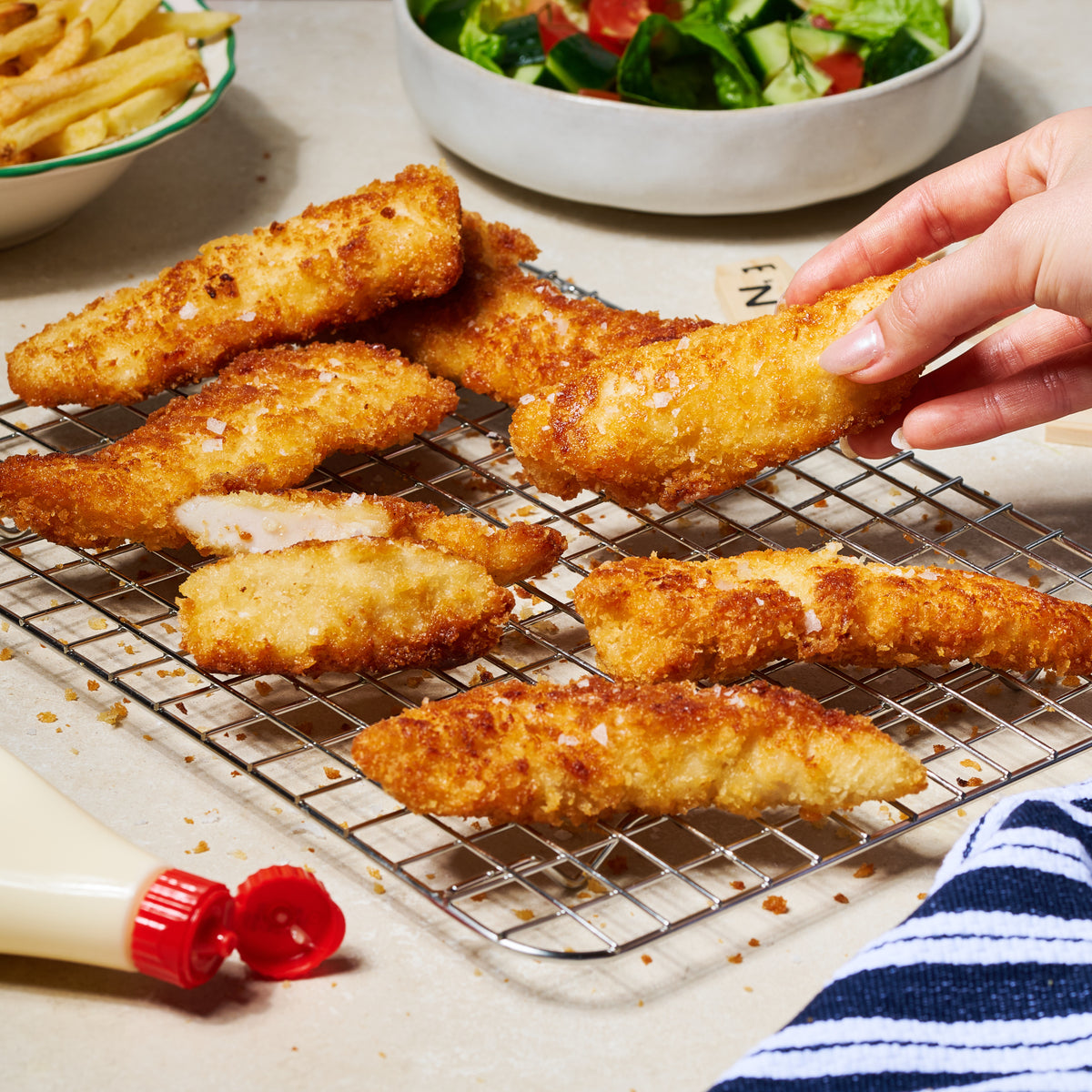 7 pieces of panko-crusted chicken tenderloins on a wire rack, served with chips and salad in the background