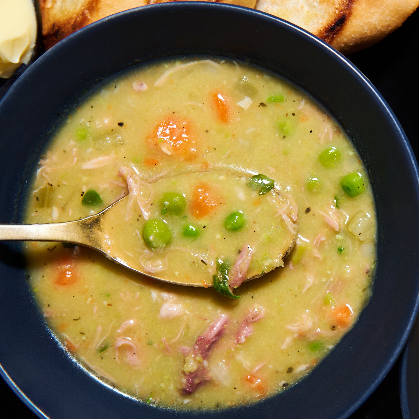 Bowl of creamy green pea and ham soup topped with peas, carrots, and tender ham, with a golden spoon lifting a mouthful.