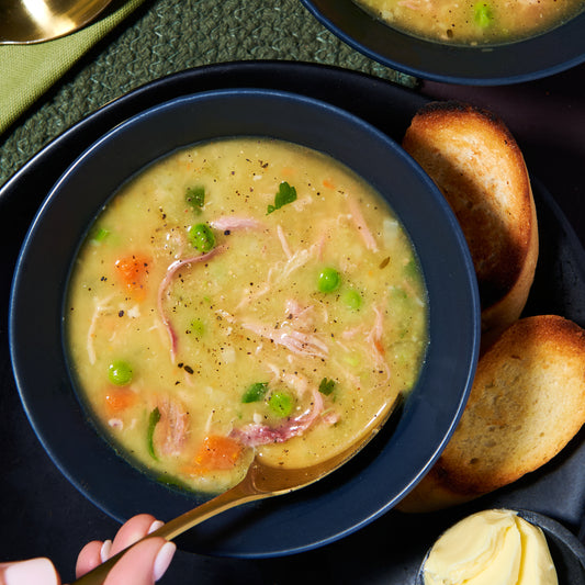 Close-up of a hearty bowl of green pea and ham soup with carrots, peas, and shredded ham, served with toasted bread on the side.