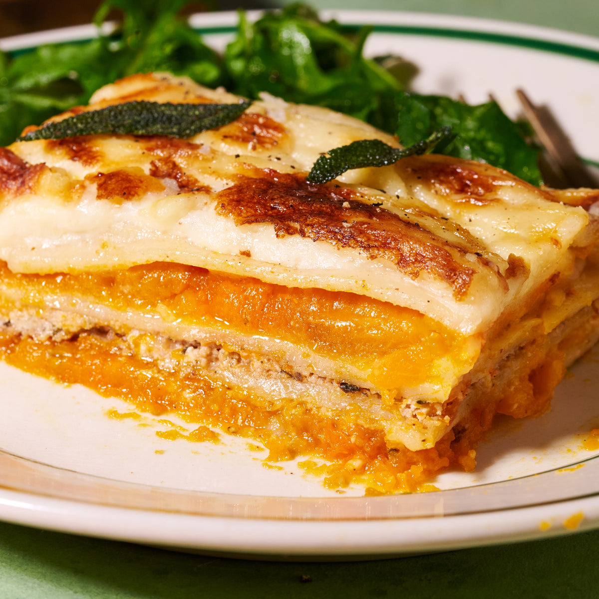 Close-up of roast pumpkin and ricotta lasagne, showing layers of pasta, roast pumpkin and creamy ricotta beneath a golden baked top, garnished with crispy sage