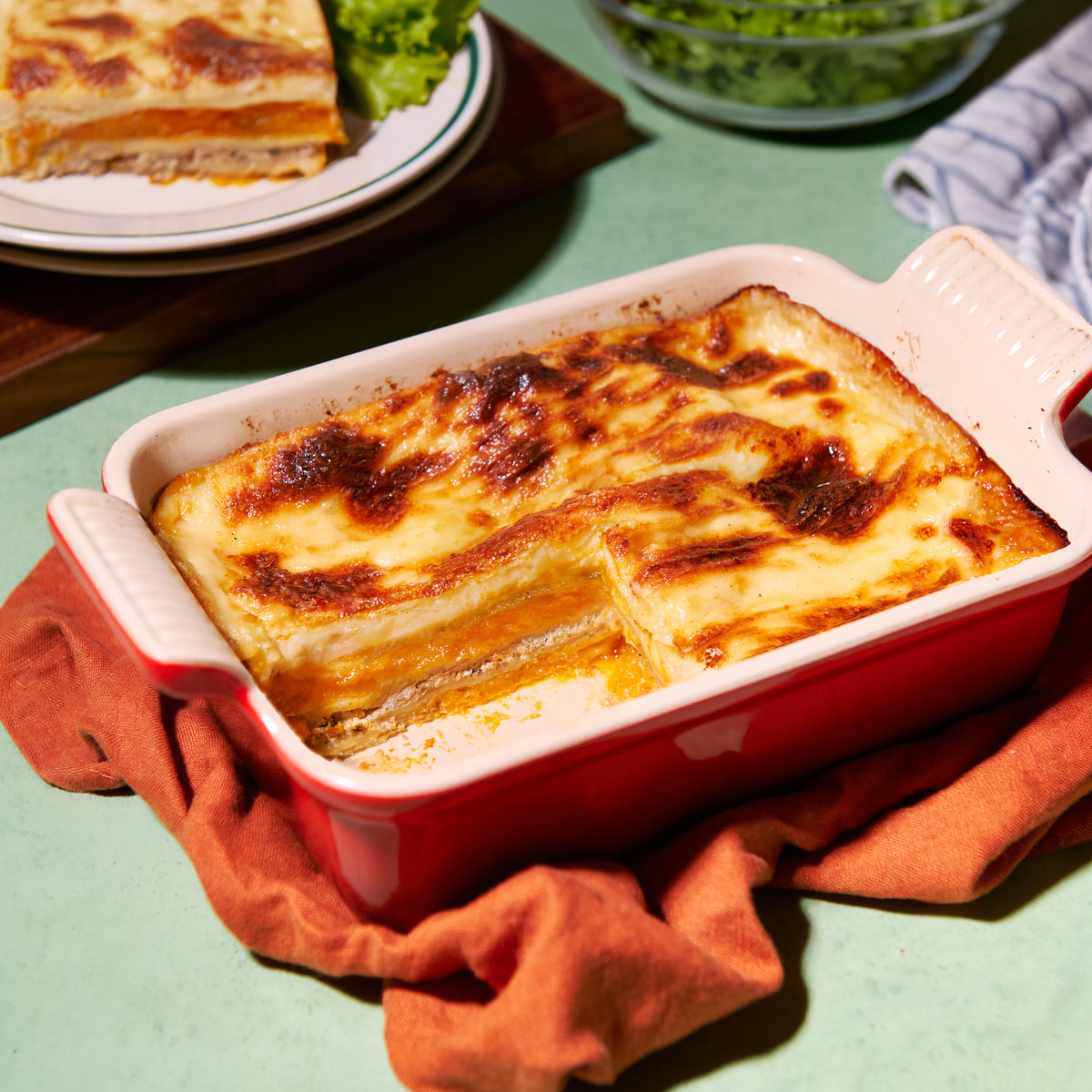 Roast pumpkin and ricotta lasagne in a baking dish, layered with golden baked cheese, with a plated slice and side salad in the background
