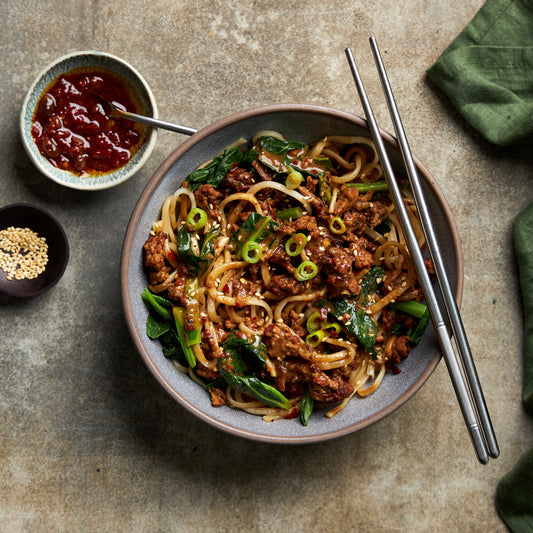 Bowl of spicy dan dan noodles with pork mince, Chinese broccoli, and shiitake mushrooms in a miso-chilli sauce, topped with spring onions.