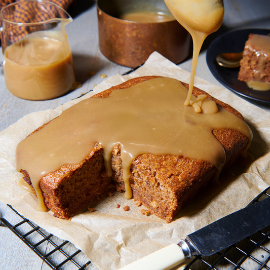 Sticky date pudding loaf on a cooling rack, generously drizzled with golden toffee sauce, with extra sauce in a jug behind.