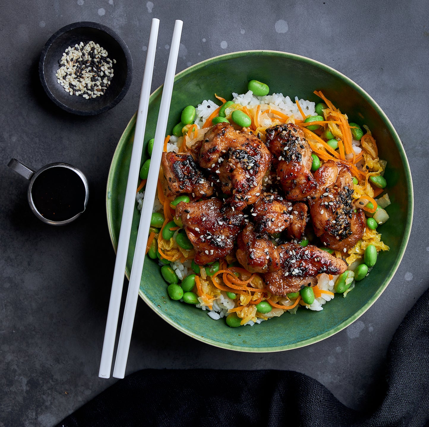 Teriyaki Chicken and Rice Bowl with glazed chicken, sushi rice, edamame & carrots, topped with sesame seeds and served with chopsticks