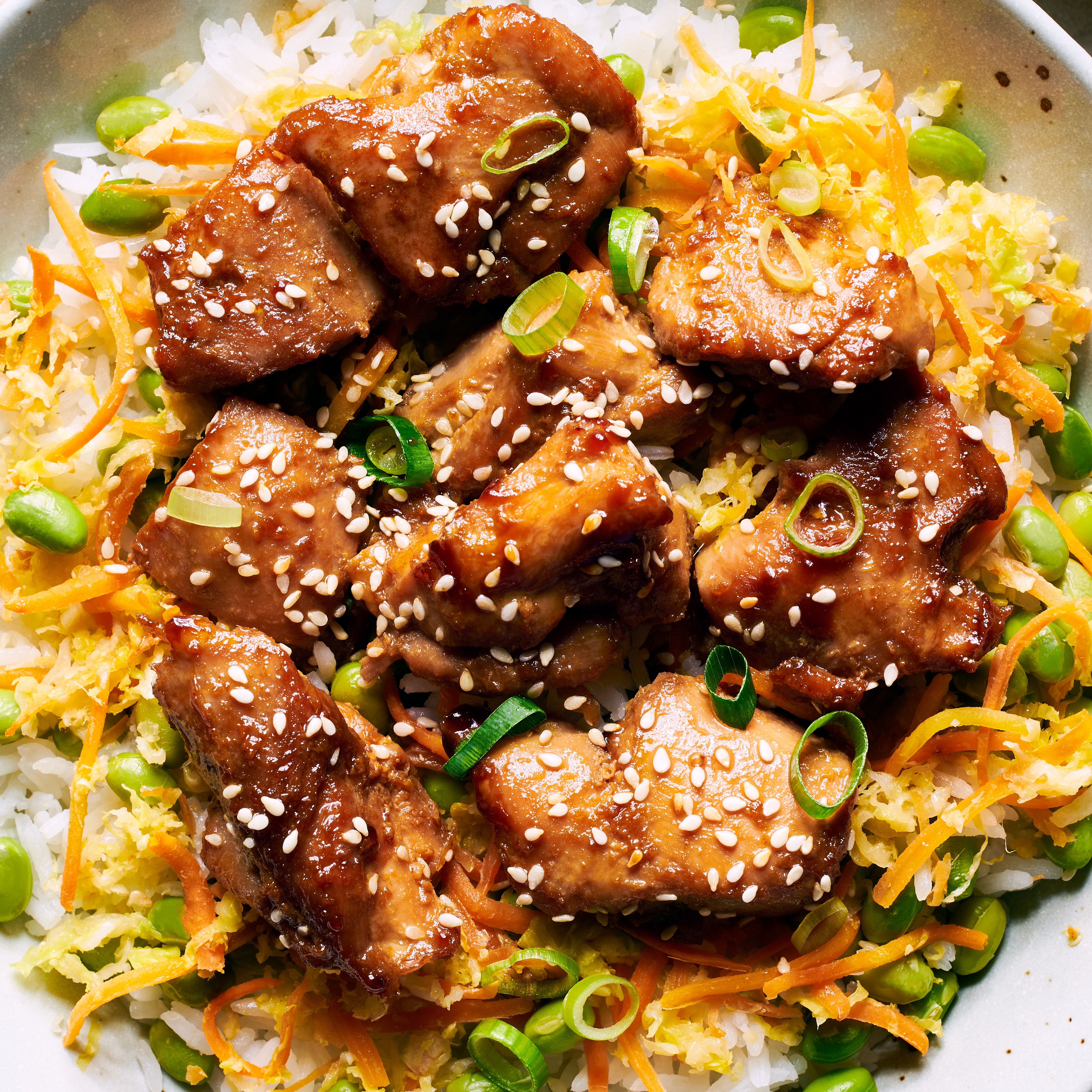 Close up of teriyaki chicken rice bowl showing juicy, glazed chicken a top a bed of sushi rice, edamame and shredded cabbage and carrots. Garnish with spring onion and roasted sesame seeds
