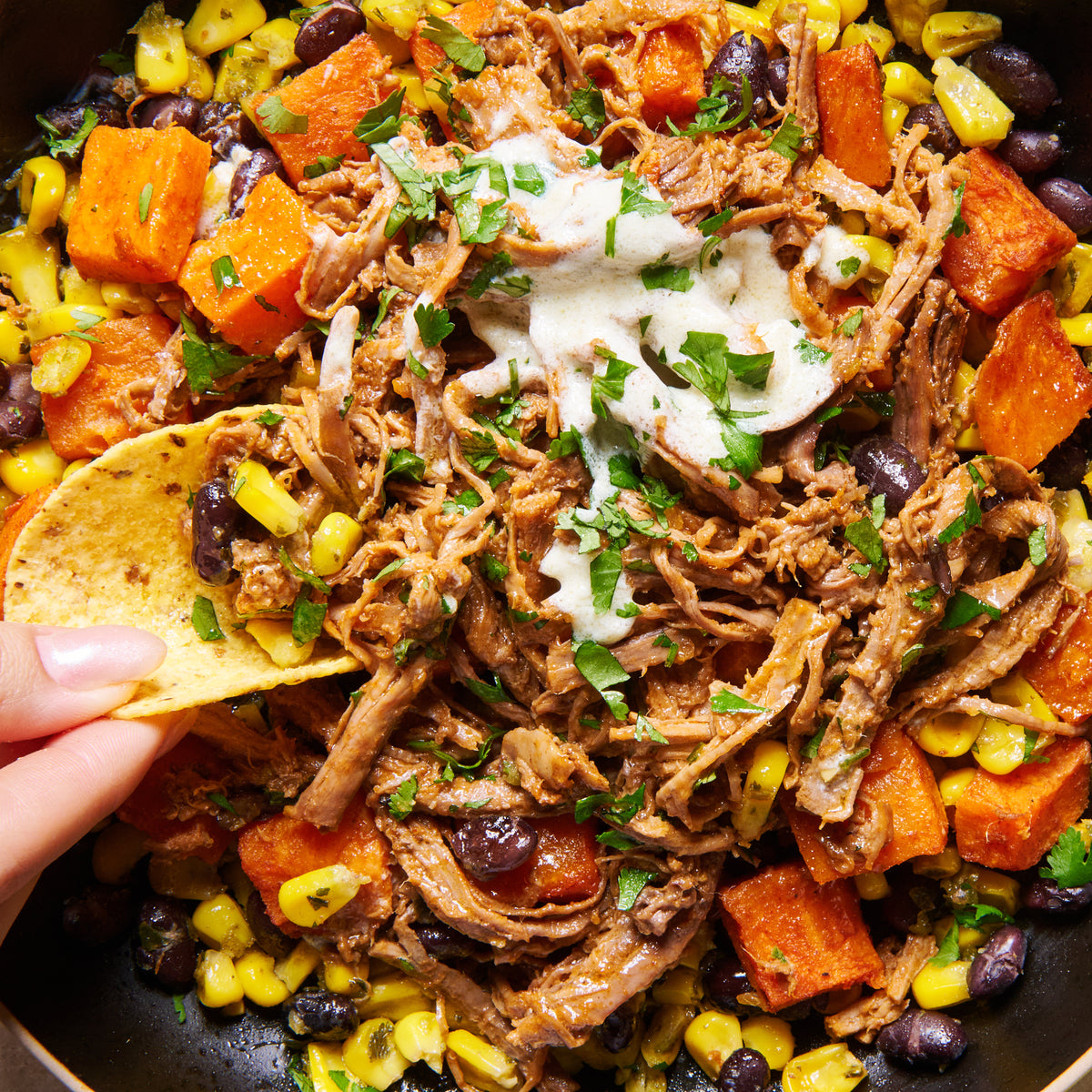 Close-up of shredded Texan beef with sweet potato, corn and black beans, drizzled with jalapeño crema and scooped with a corn chip