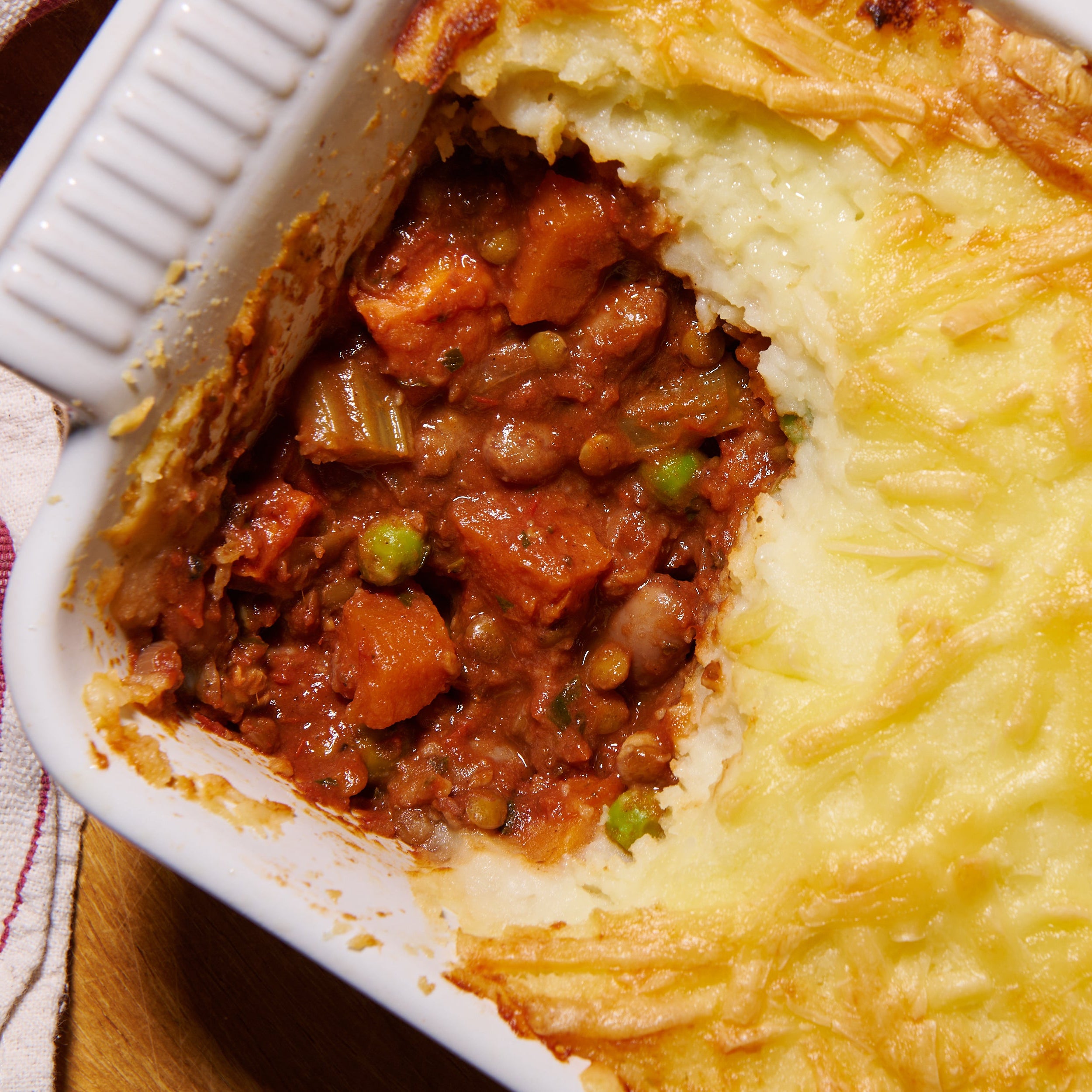 Close-up of vegetable cottage pie, showing roasted carrots, Puy lentils, borlotti beans and green peas beneath a layer of golden cheesy mash