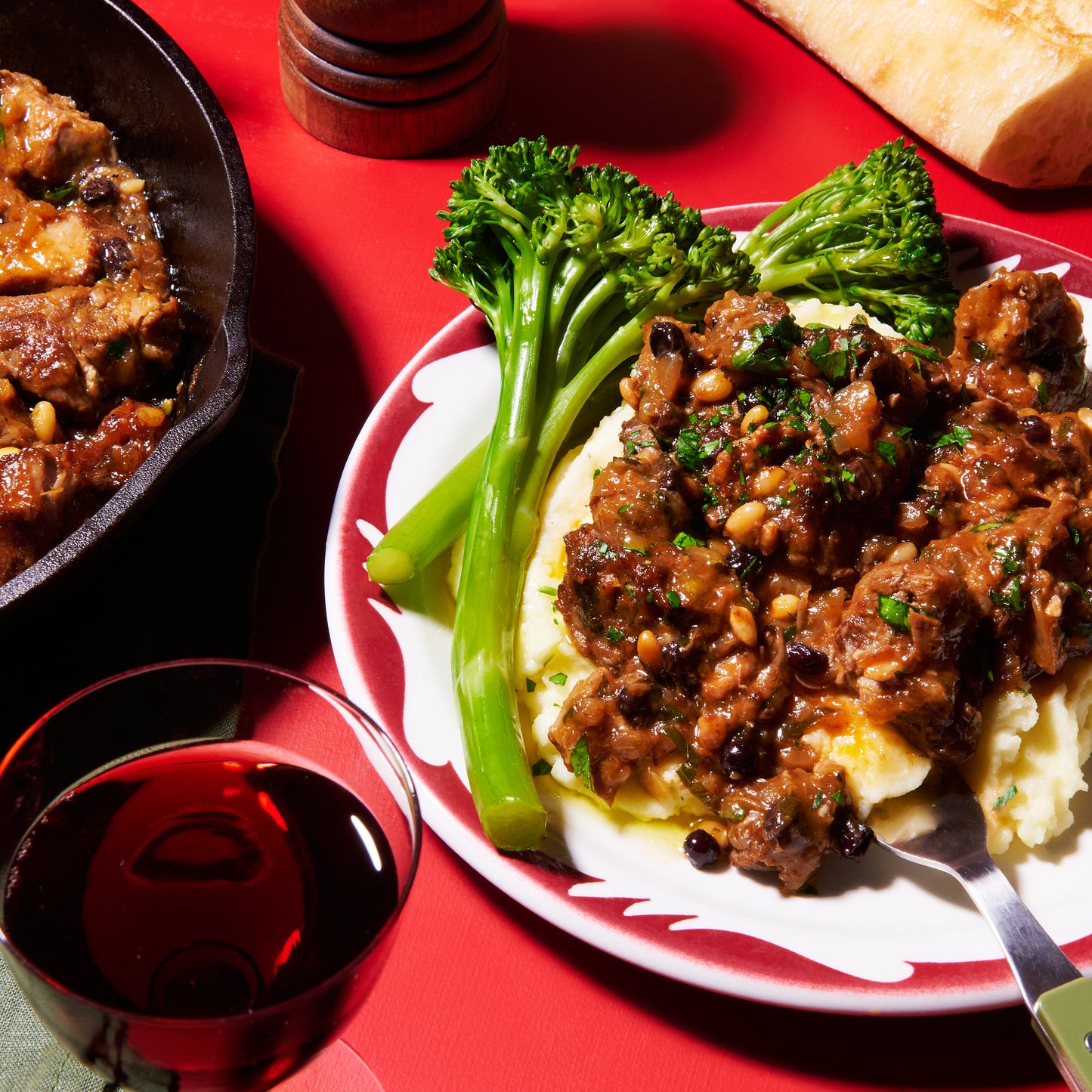 Plate of agrodolce lamb served over mashed potatoes with broccolini and a glass of red wine.