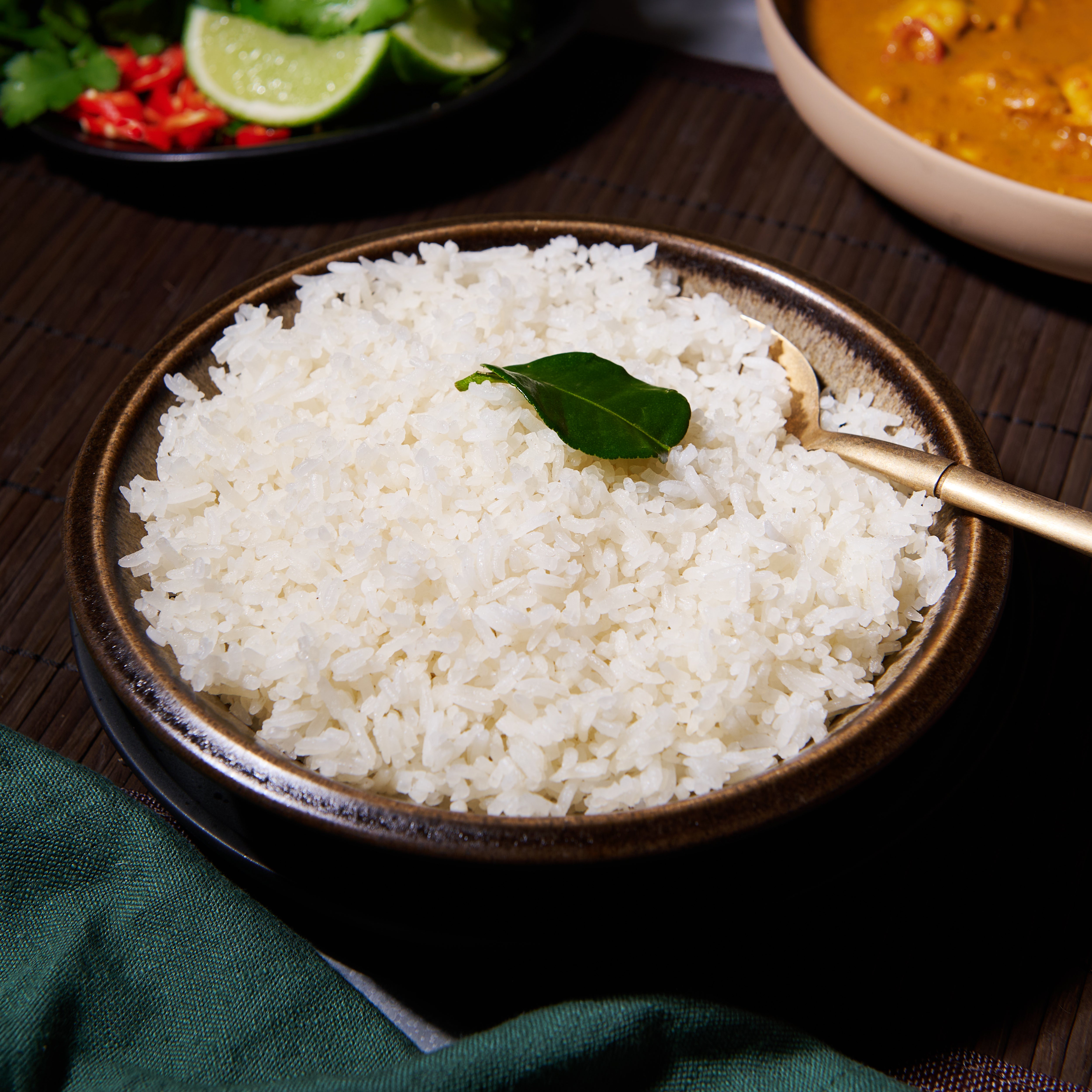 Steamed coconut white rice in a dark bowl garnished with a keffir lime leaf, served with a curry and fresh chilli and lime on the side