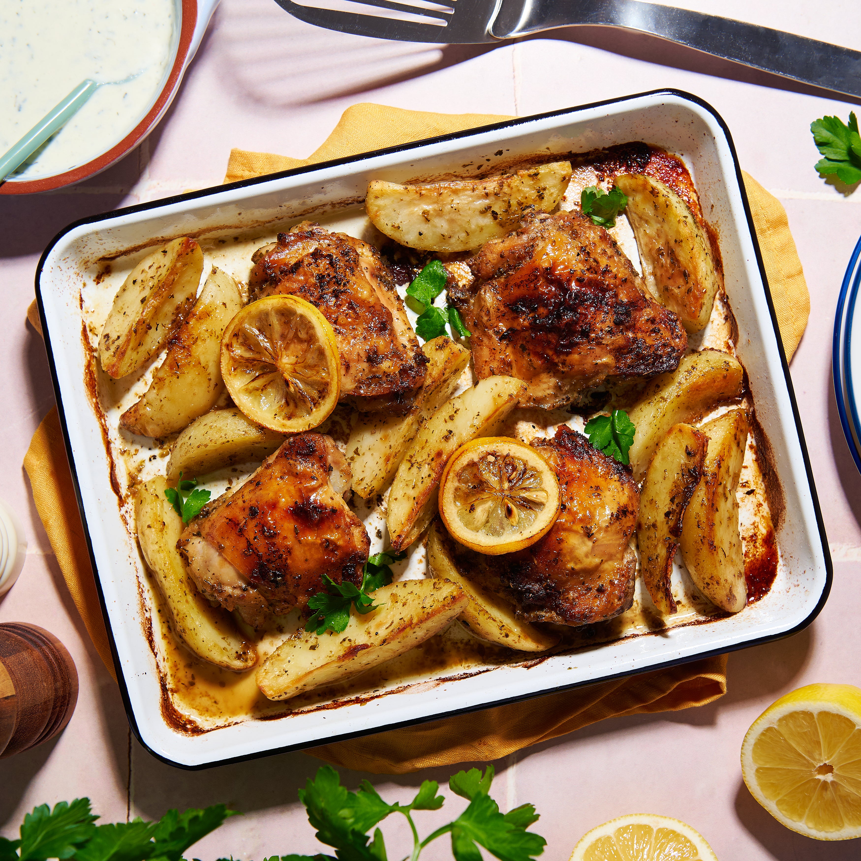 Golden roasted chicken thighs with potato wedges, lemon slices, and oregano in a rustic Greek traybake.