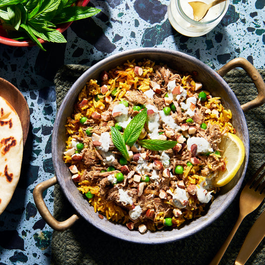 A shallow metal bowl filled with golden lamb biryani, topped with slow-cooked lamb, peas, toasted nuts, fresh mint leaves, and drizzles of yoghurt.