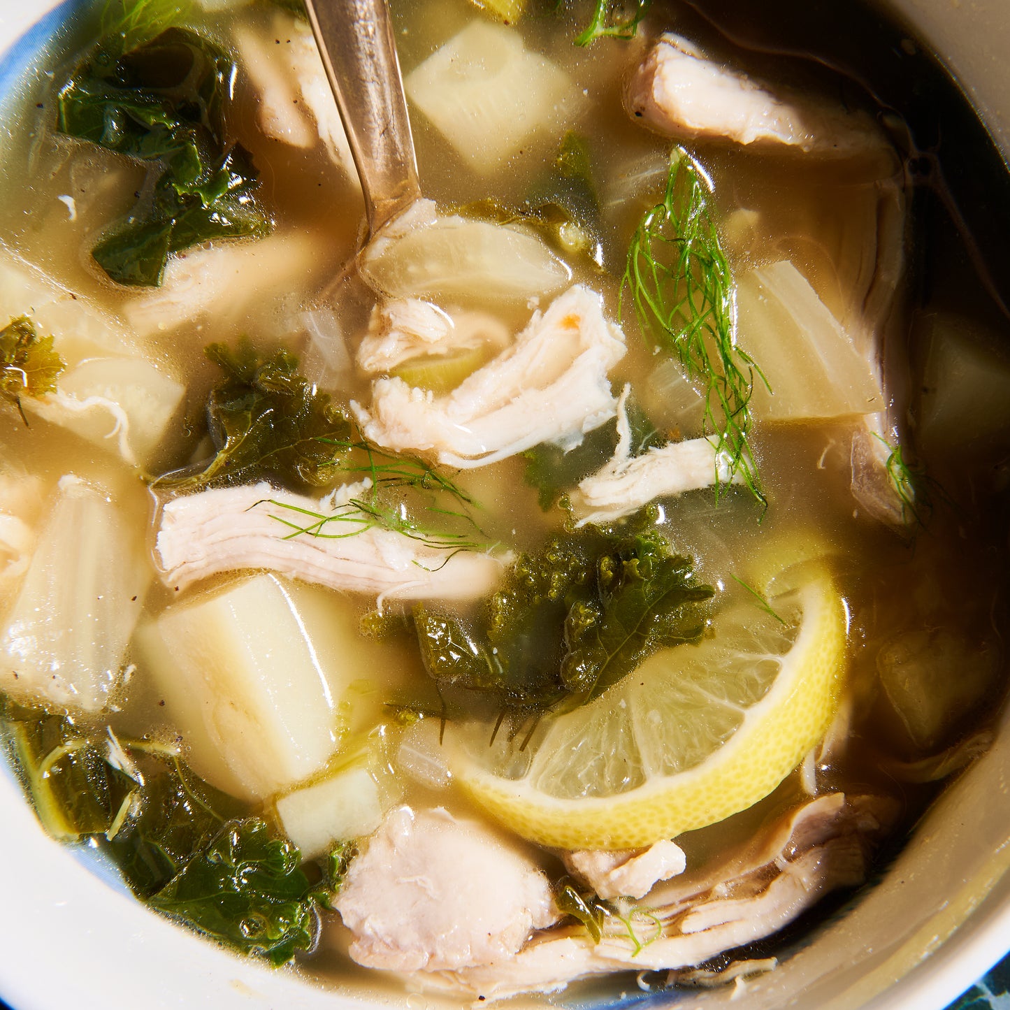 Close-up of Lemony Chicken Soup showing shredded chicken, lemon slice, kale, fennel, and diced potatoes in broth with a spoon.