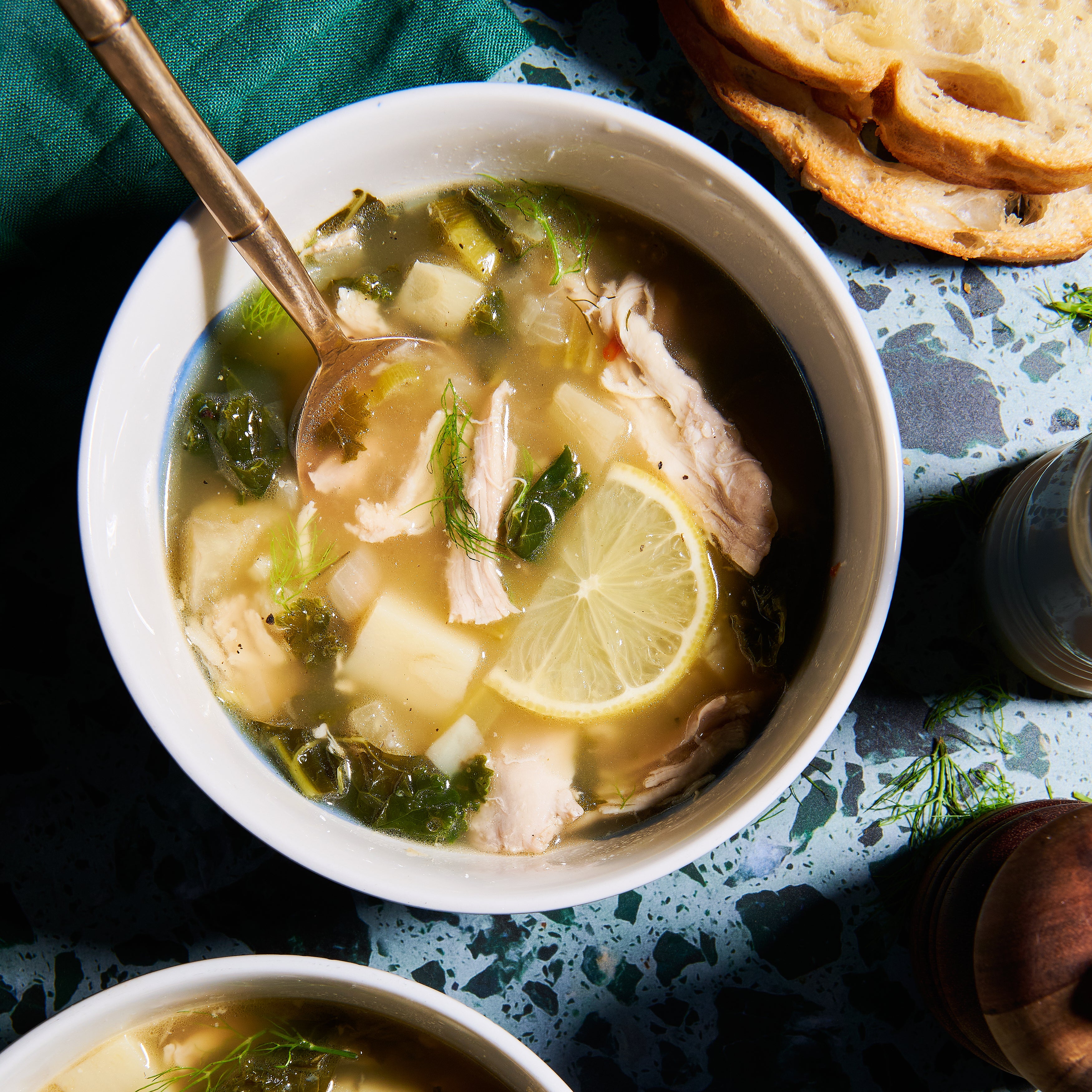 Overhead view of a bowl of Lemony Chicken Soup with lemon, chicken, and greens, served with crusty bread on the side.