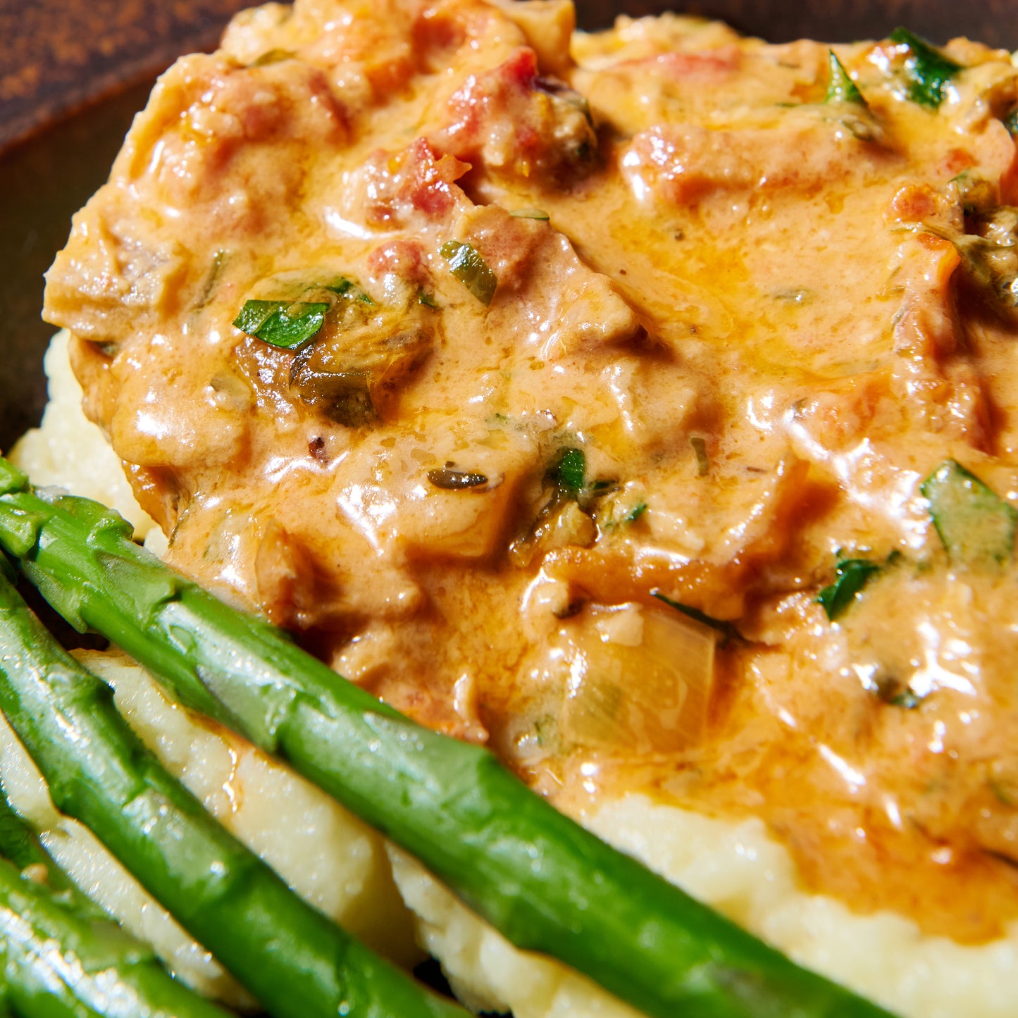 Close-up of creamy sun-dried tomato sauce with tender chicken pieces served alongside green asparagus and mashed potato.