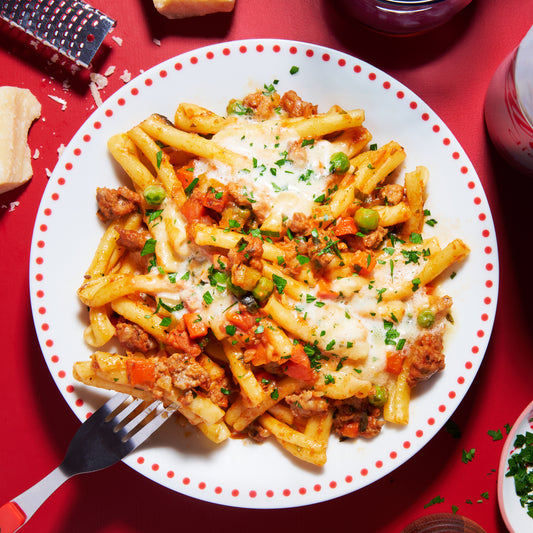 Bowl of casarecce pasta with Italian sausage, pancetta, peas and parmesan cream sauce on a red background.