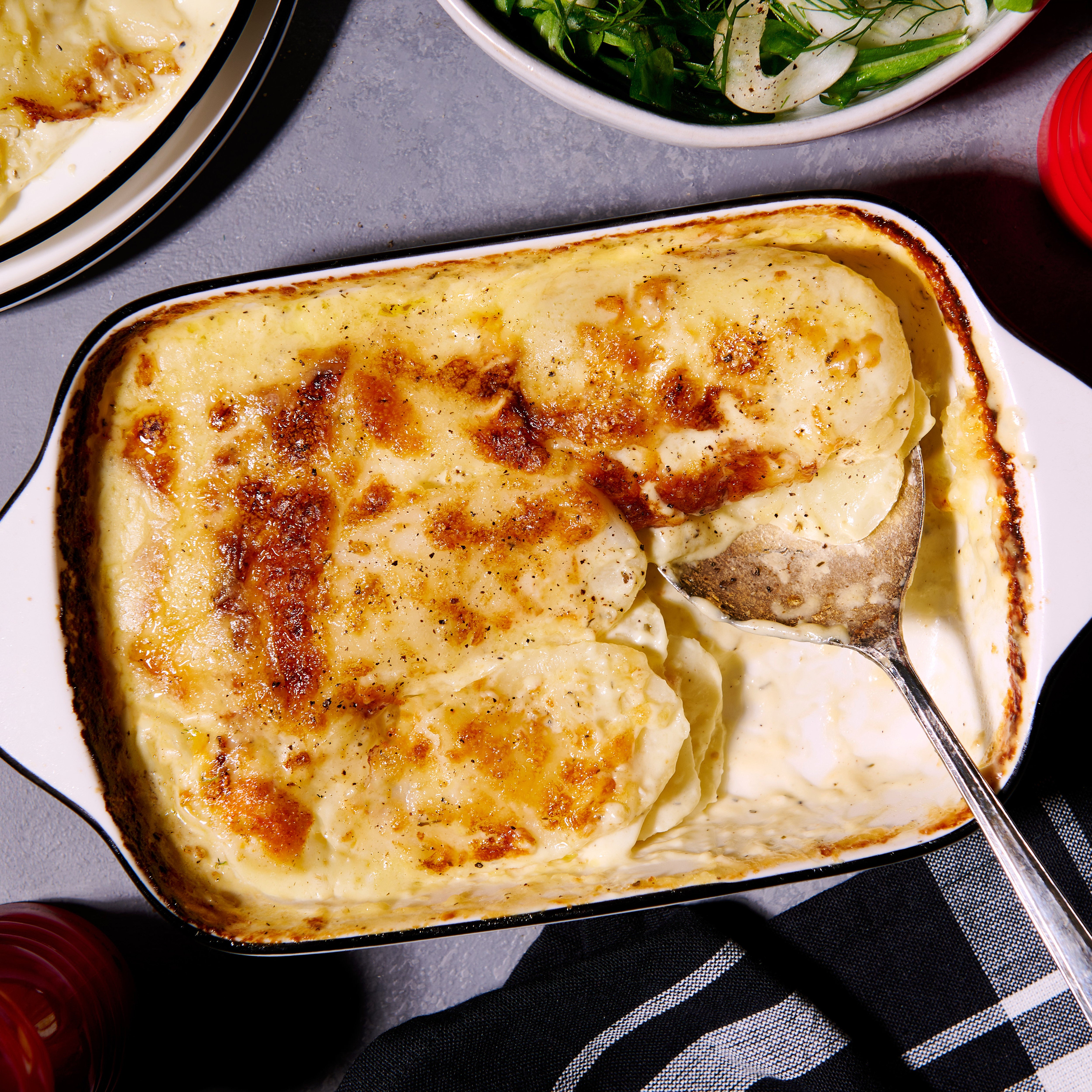 Potato Gratin baked with thinly sliced potatoes in garlic-Parmesan cream, topped with golden Gruyere cheese, served in a baking dish with a portion taken out. Fennel and rocket salad on the side