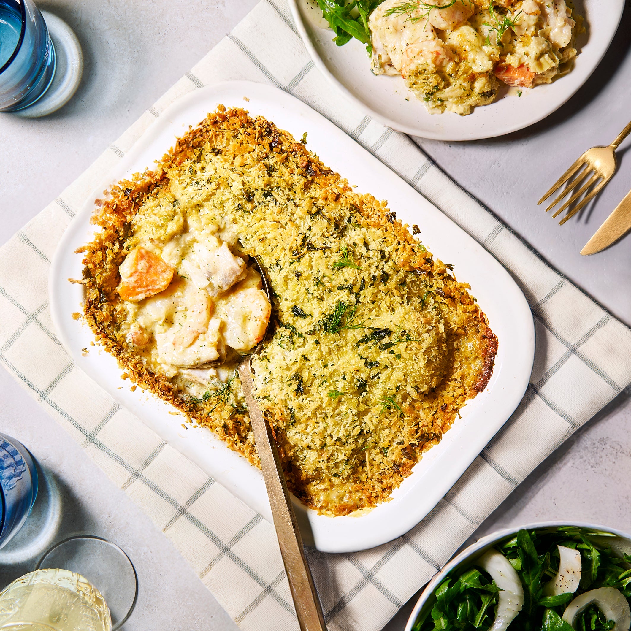 Overhead view of Prawn, Leek & Barramundi Gratin in a white baking dish with golden crumb topping, served with salad on the side.