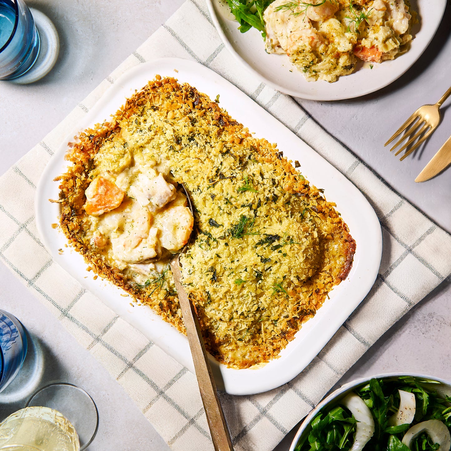 Overhead view of Prawn, Leek & Barramundi Gratin in a white baking dish with golden crumb topping, served with salad on the side.