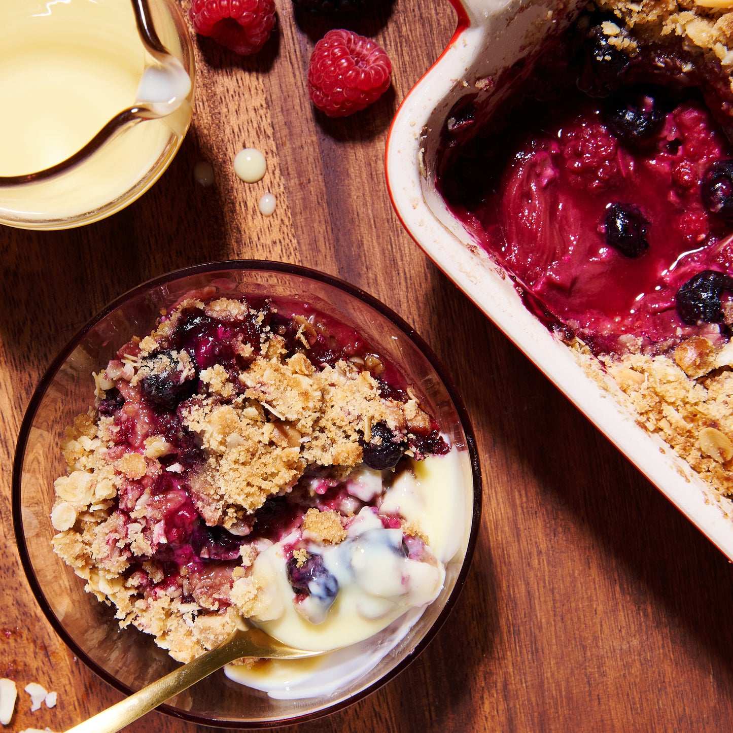 A serving of rhubarb and berry crumble in a glass bowl, topped with custard and surrounded by fresh berries.