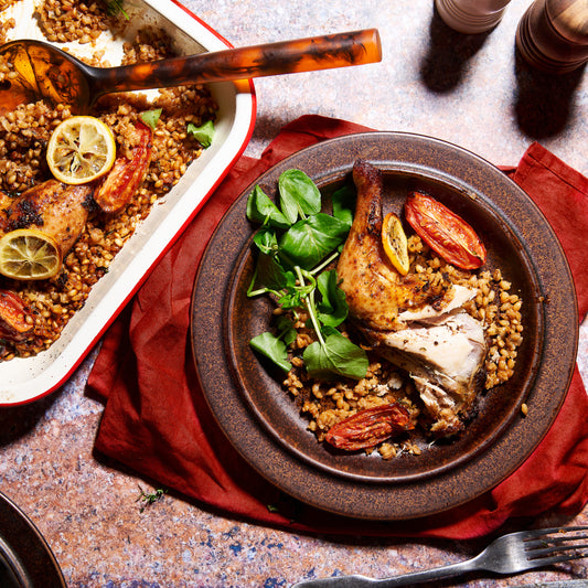 Plate of Mediterranean Roast Chicken with pearl barley risotto, roast tomatoes, lemon slices, and greens, next to a serving tray.