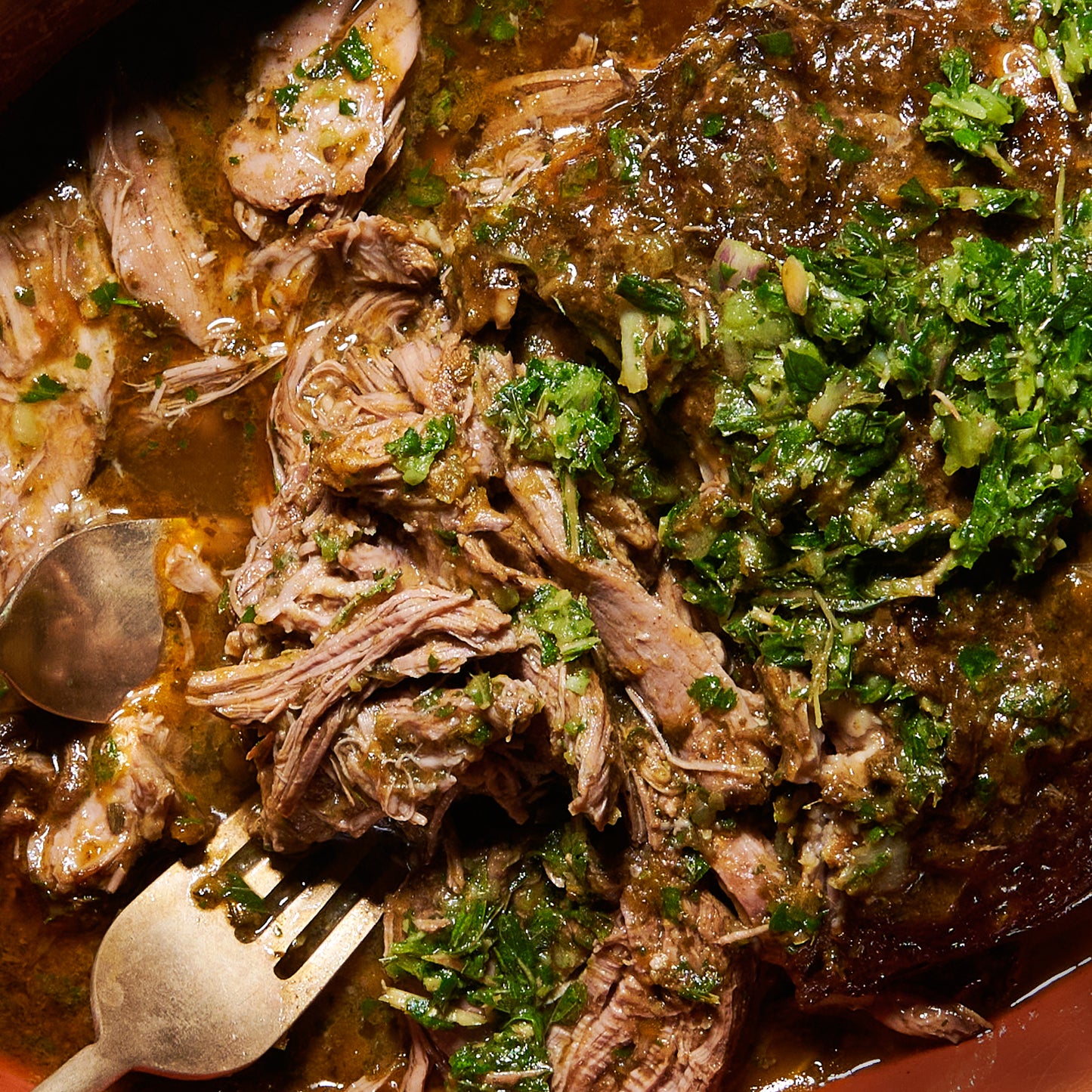 Close-up of slow-cooked lamb shredded in pan juices, garnished with fresh mint salsa verde.