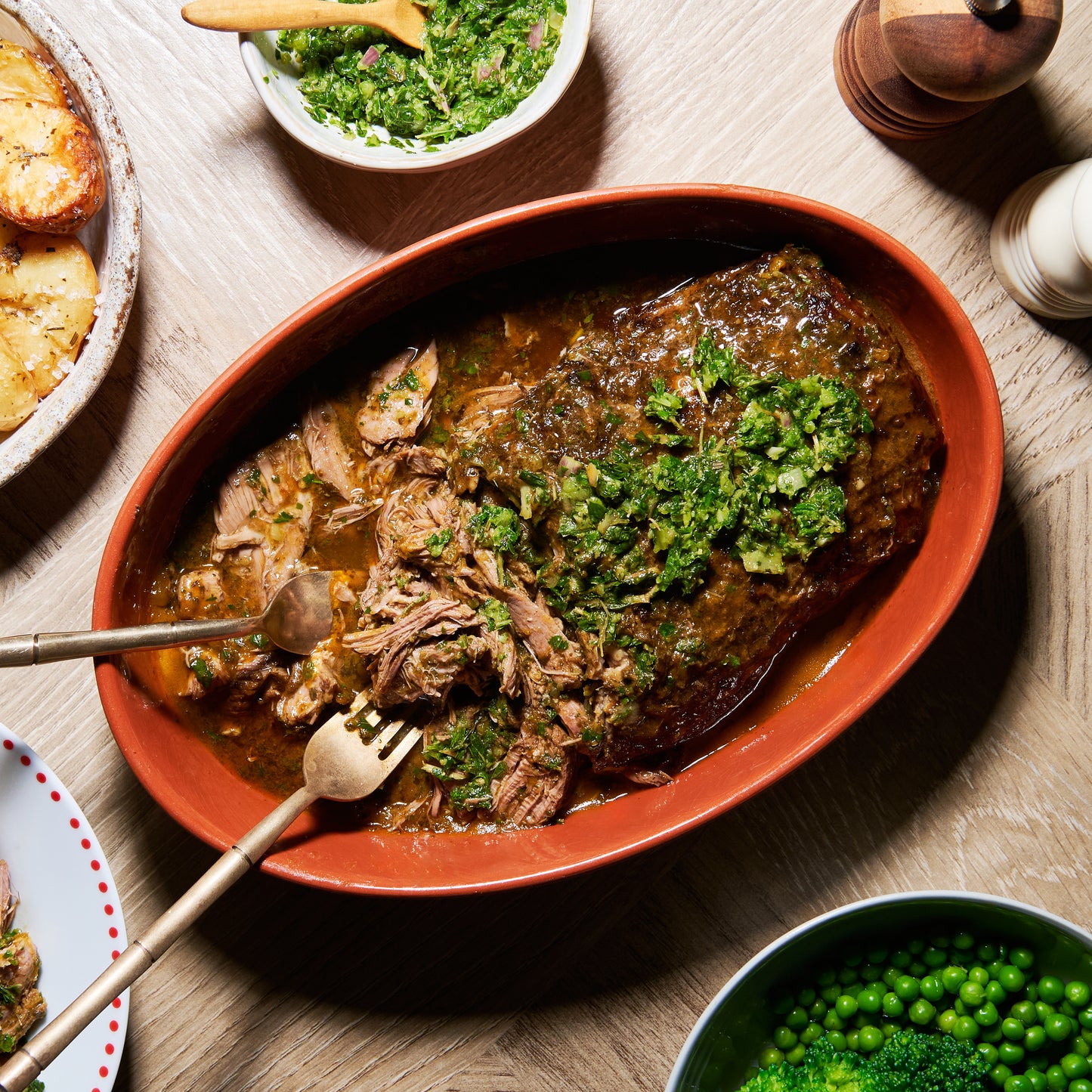 Platter of herby slow-cooked boneless leg of lamb topped with mint salsa verde, served with roasted potatoes and peas.