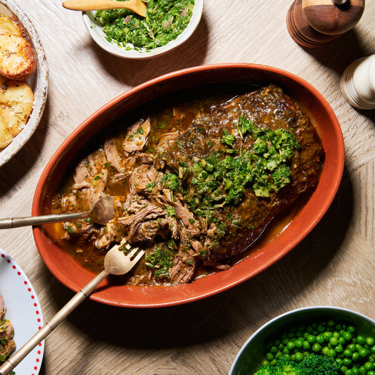 Platter of herby slow-cooked boneless leg of lamb topped with mint salsa verde, served with roasted potatoes and peas.