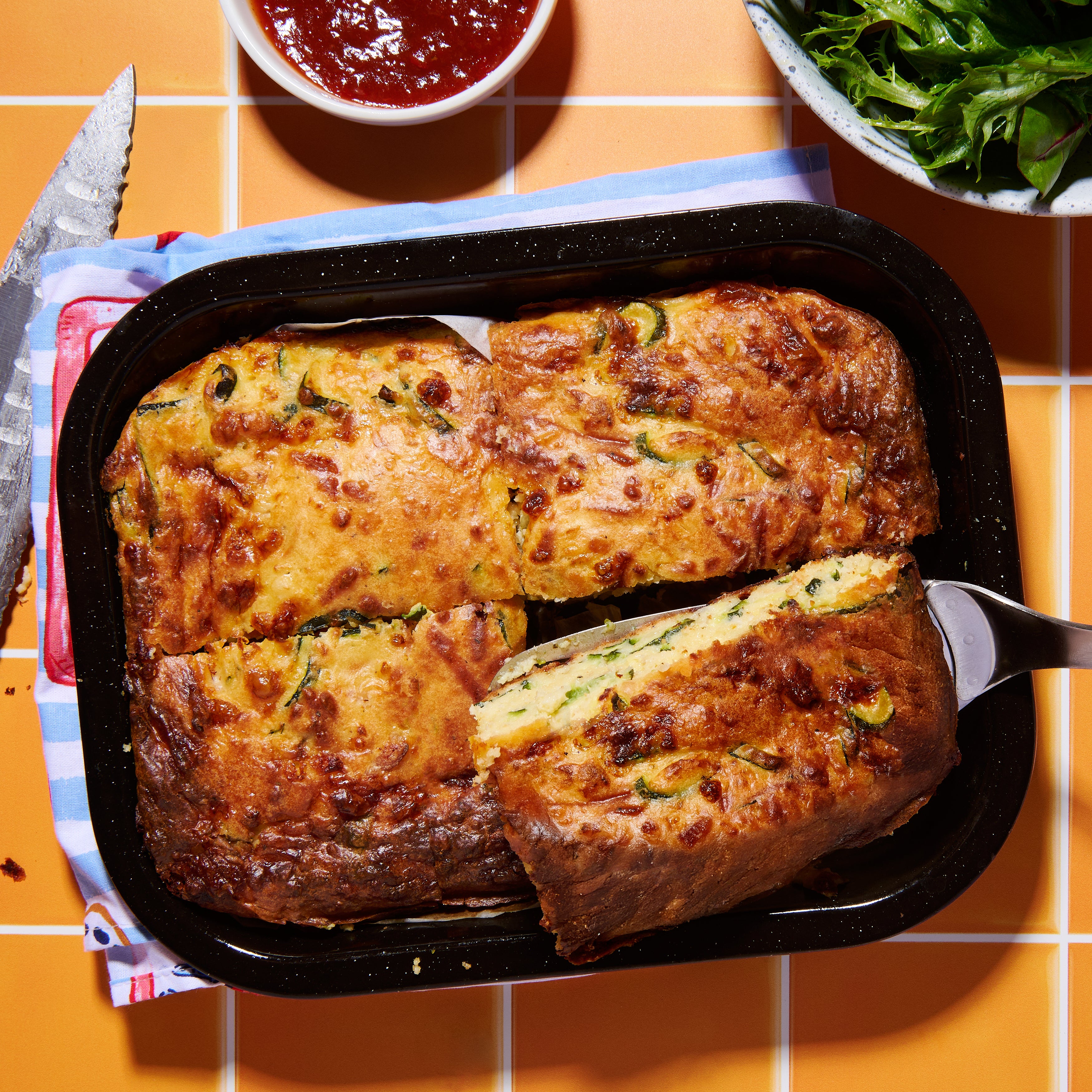 Golden-baked zucchini and pancetta slice in a tray, cut into portions and ready to serve with salad and sauce on the side.