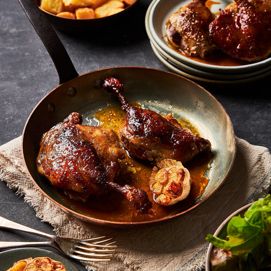 Golden, crisp-skinned duck confit glazed with citrus and maple syrup in a rustic pan, served with roasted garlic and greens.