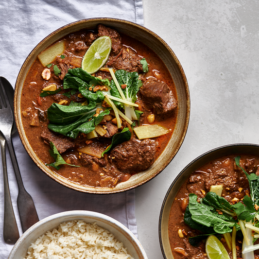 Bowl of Thai red beef curry with bamboo shoots, peanuts, lime wedge, and fresh Asian greens served with steamed rice.
